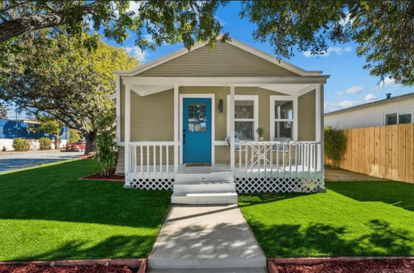 Small olive green house featuring a bright blue door and a white front porch.