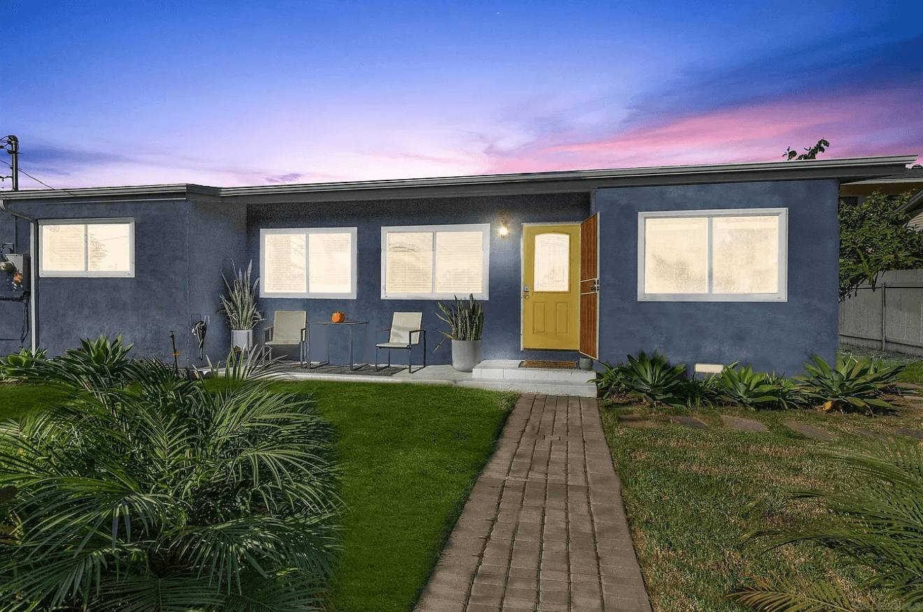 Blue stucco house with a yellow door, brick walkway, and lush garden at sunset.