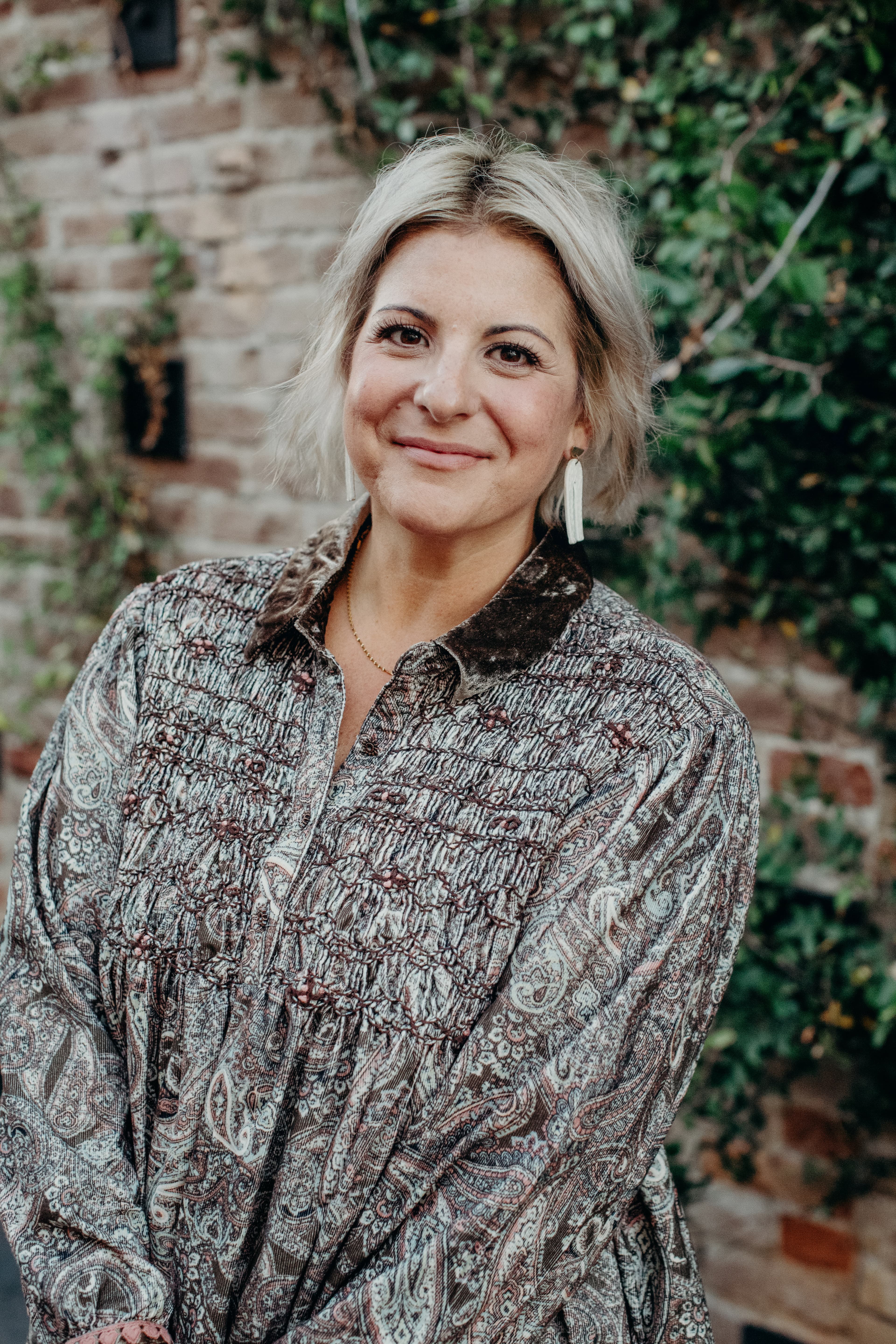 Smiling blonde woman in a paisley blouse against a brick wall with green vines.
